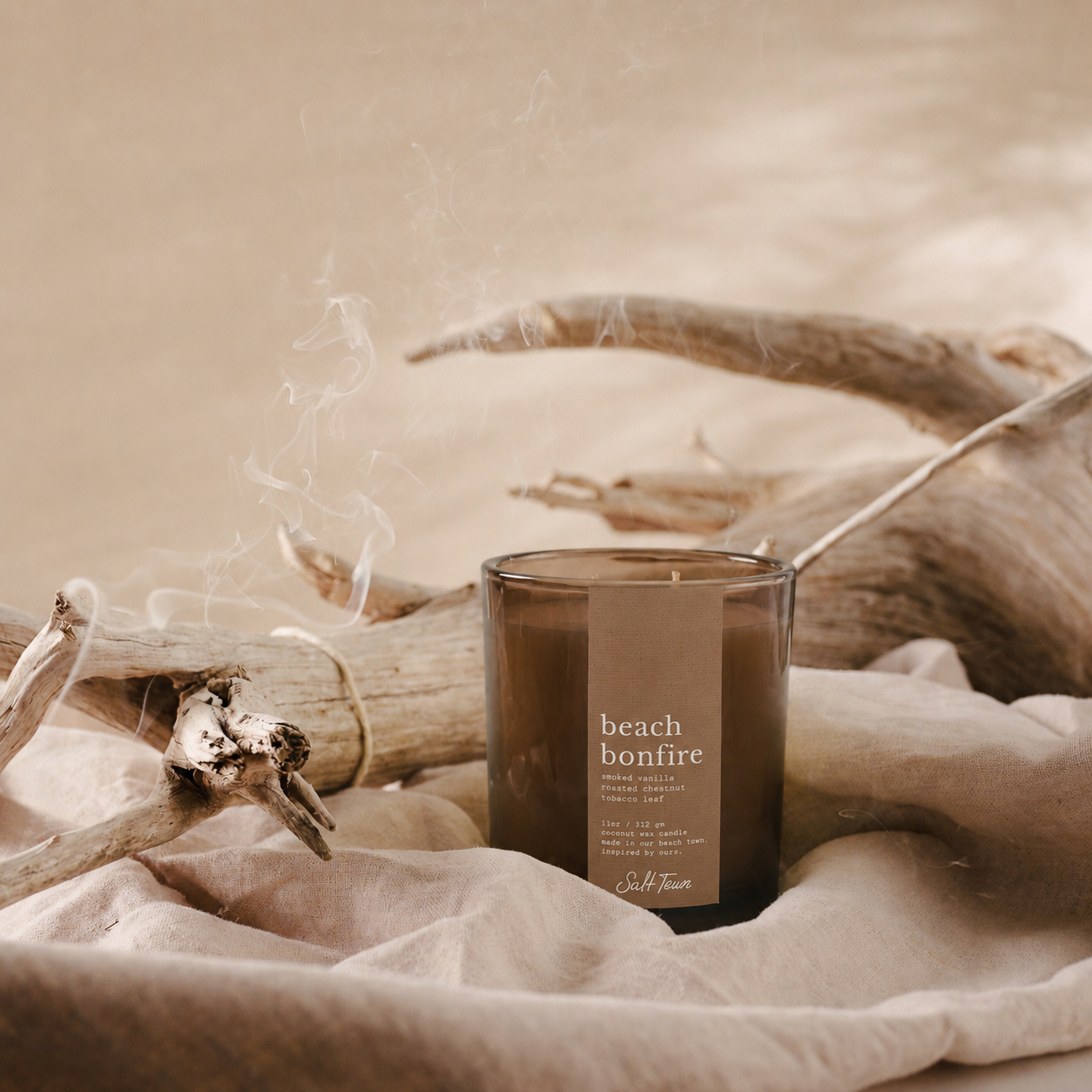 A smoky amber glass candle jar labeled 'beach bonfire' by Salt Town, styled on beige fabric with driftwood branches in a warm, neutral-toned setting.
