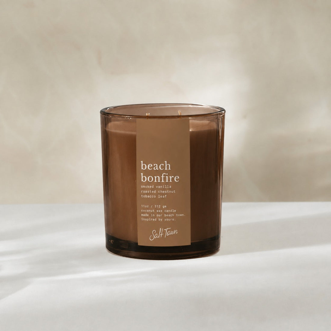 A beach bonfire scented candle in a translucent amber glass jar with a brown paper label, set against a warm beige background.