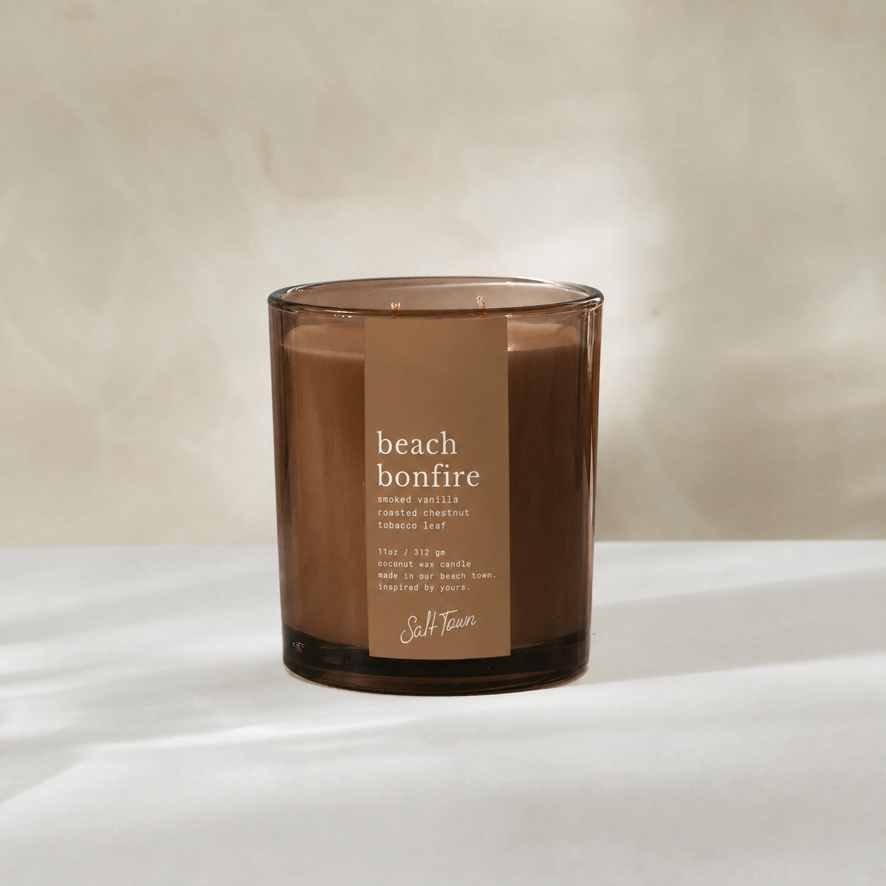 A beach bonfire scented candle in a translucent amber glass jar with a brown paper label, set against a warm beige background.