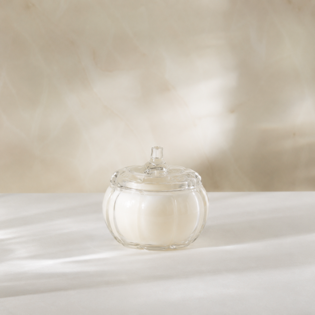 A clear glass pumpkin-shaped candle with a lid and white wax, set on a white draped surface against a warm beige background.
